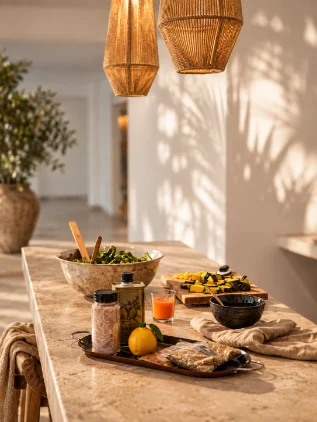 A stone counter with a salad bowl, olive oil, pink salt, a lemon, a glass of orange juice and squash on a cutting board, under woven pendant lights.